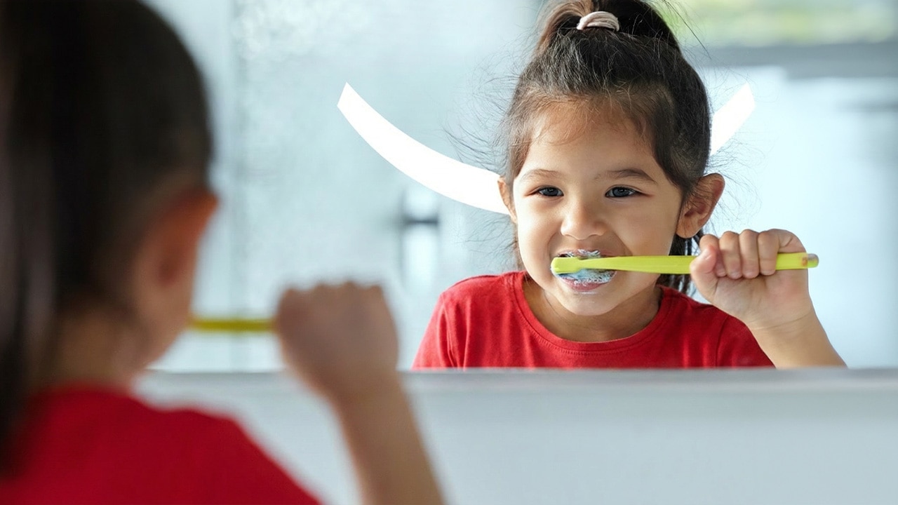 Une petite fille se brossant les dents tout en regardant son reflet dans le miroir, avec le texte « Dentifrice au fluor pour enfants ».