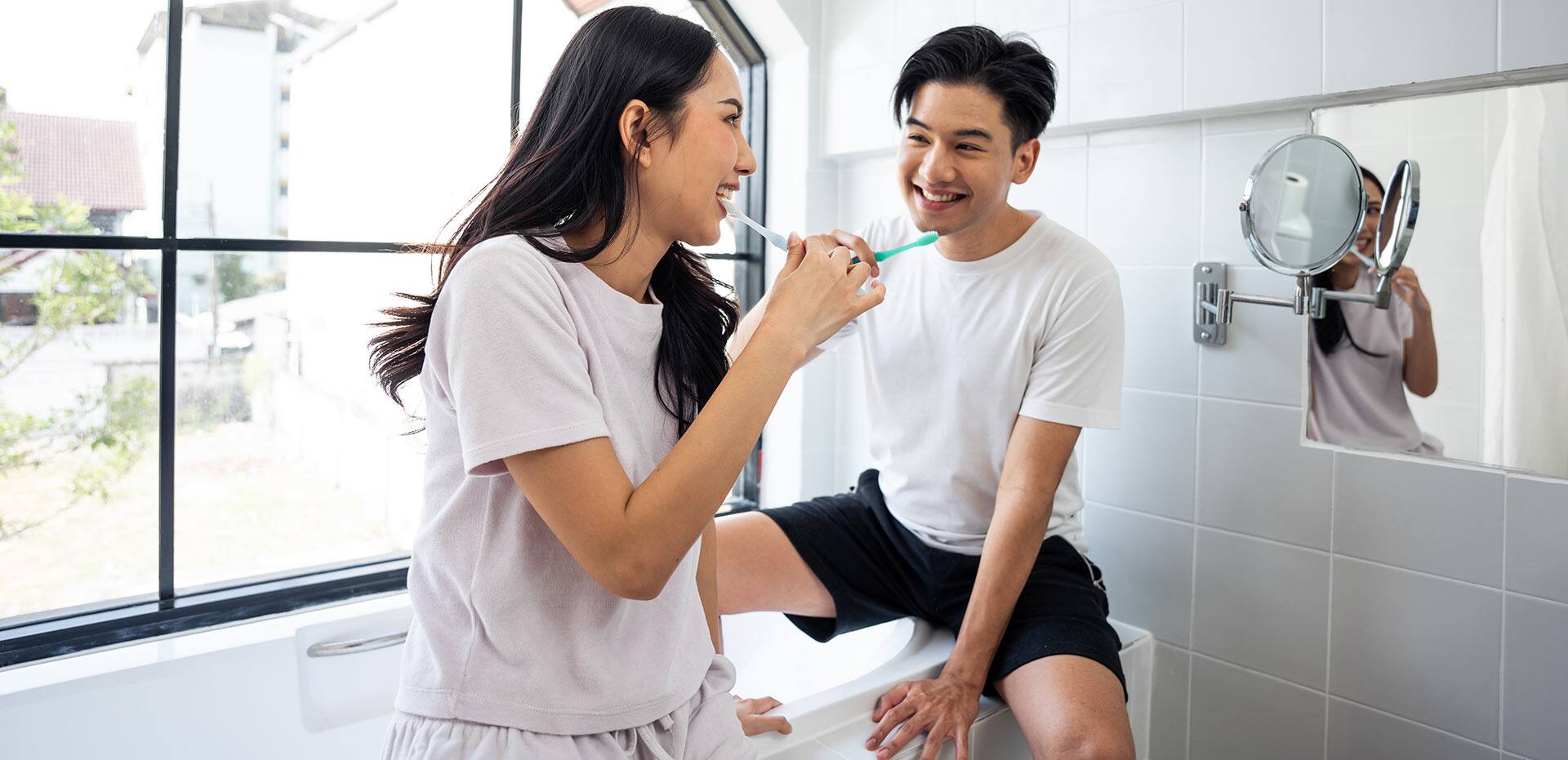 Asian new marriage couple brushing teeth together in bathroom at home. Attractive young man and woman feeling happy and relax, enjoy holiday honeymoon anniversary day then looking each other in house.