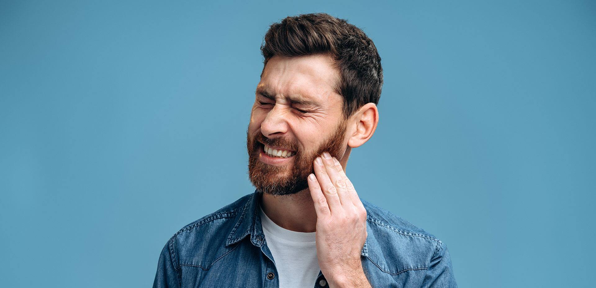 Dental problems. Portrait of unhealthy man pressing sore cheek, suffering acute toothache, periodontal disease, cavities or jaw pain. Indoor studio shot isolated on blue background
