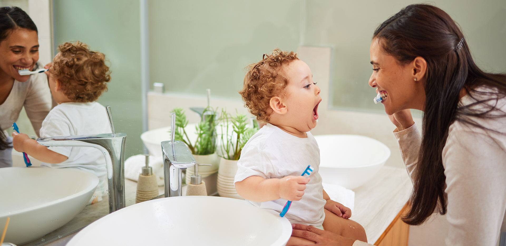 Mom teaching baby to brush its teeth, on the bathroom counter in home and a clean smile on her face. Healthy oral hygiene for kid means using child friendly toothpaste, toothbrush and dental routine