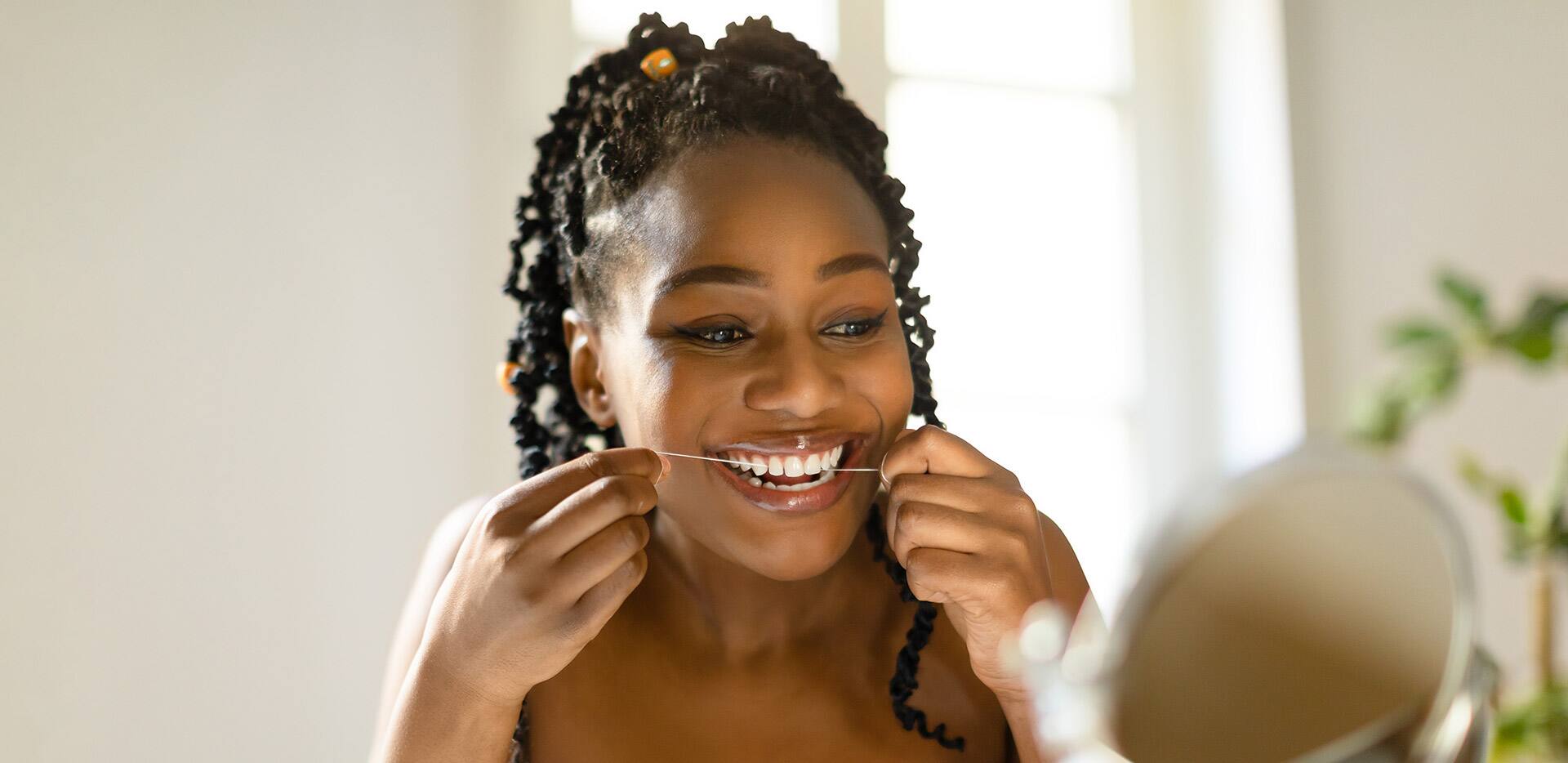 Attractive black woman looking at mirror and using dental floss, cleaning her white teeth, sitting at vanity table